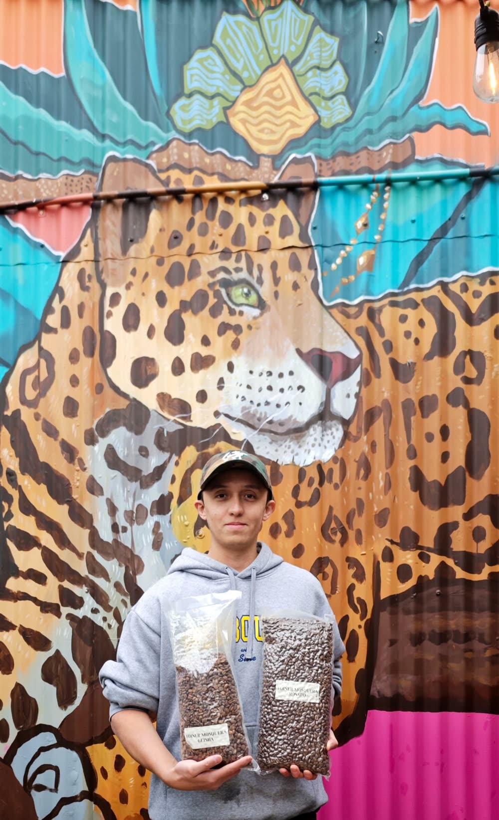 Nicolás Bonilla Moreno, founder of Kitsu Coffee, holding two bags of green coffee in front of a jaguar mural